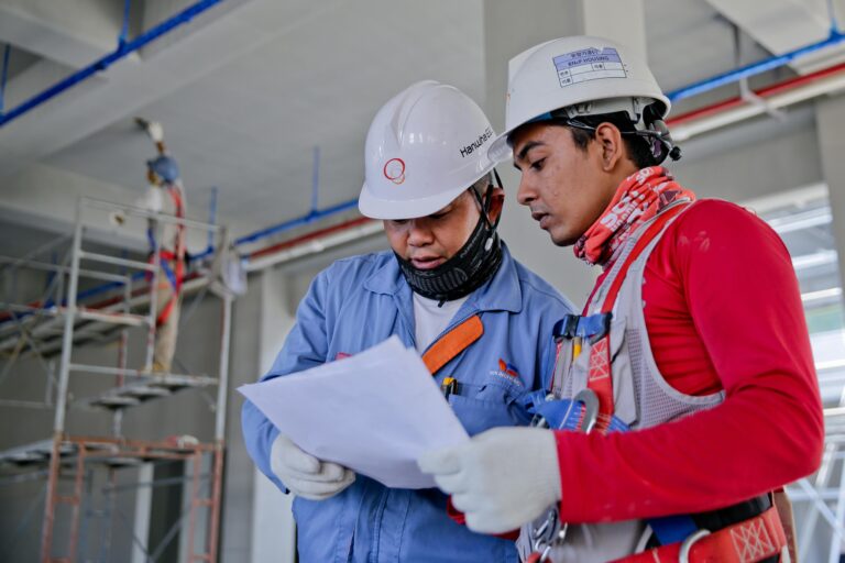 Two engineers in safety helmets reviewing construction plans at a worksite.